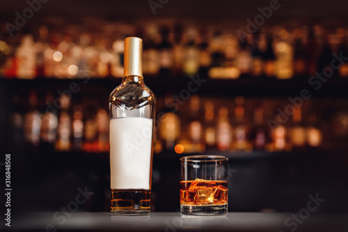Fotografía Glass of whiskey bottle with ice stands on bar counter, dark brown background