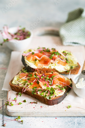 Toast with avocado cream and smoked salmon on the white wooden board. Smoked salmon, cream cheese and pesto toast sandwiches with radish sprouts