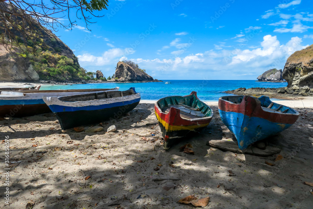 A row of colorful boats parked on a shore of idyllic Koka Beach in Flores, Indonesia. The boats are lined up under the trees, in the shade. There are some cliffs in the back. Hidden gem of Indonesia. A row of colorful boats parked on a shore of idyllic Koka Beach in Flores, Indonesia. The boats are lined up under the trees, in the shade. There are some cliffs in the back. Hidden gem of Indonesia.