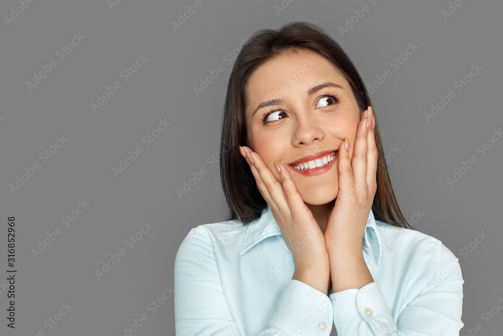 Freestyle. Young woman standing isolated on grey holding face looking aside excited close-up Freestyle. Young woman standing isolated on grey holding face looking aside excited close-up