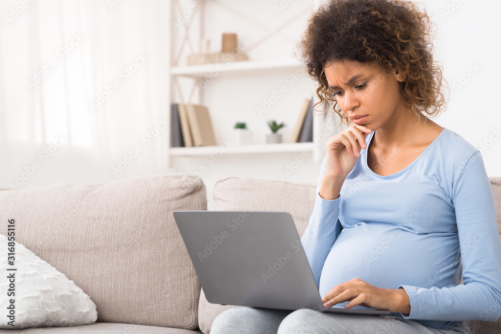 Confused afro expectant woman using laptop, buying goods for newborn Confused afro expectant woman using laptop, buying goods for newborn