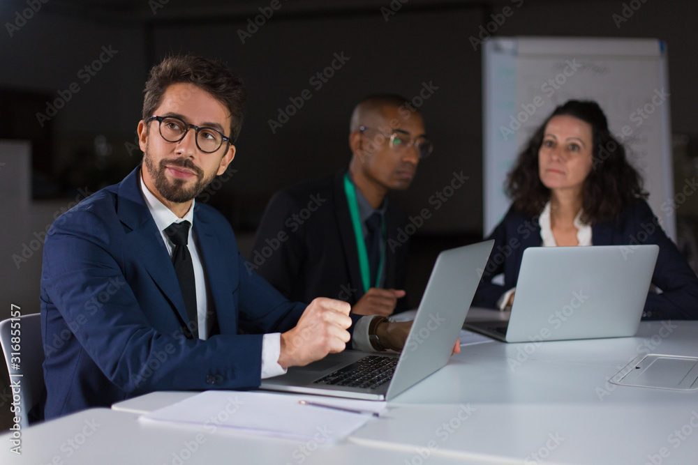 Thoughtful young man in eyeglasses looking aside. Group of confident young people sitting at table and working with laptops. Teamwork, working late concept Thoughtful young man in eyeglasses looking aside. Group of confident young people sitting at table and working with laptops. Teamwork, working late concept