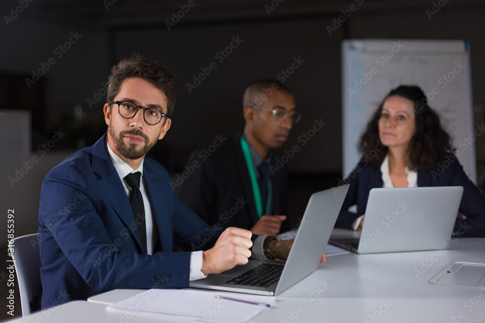 Thoughtful young man in eyeglasses looking at camera. Group of confident young people sitting at table and working with laptops. Teamwork, working late concept Thoughtful young man in eyeglasses looking at camera. Group of confident young people sitting at table and working with laptops. Teamwork, working late concept