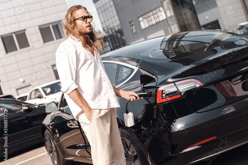 Transportation. Young man with long hair in sunglasses traveling by electric car stop at chraging station holding charging plug looking aside pensive © Viktoriia