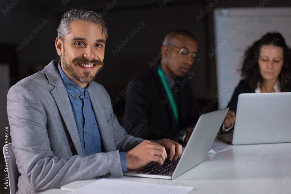 Smiling handsome manager sitting at table and looking at camera. Cheerful young man working with laptop in office at night. Business, working late concept