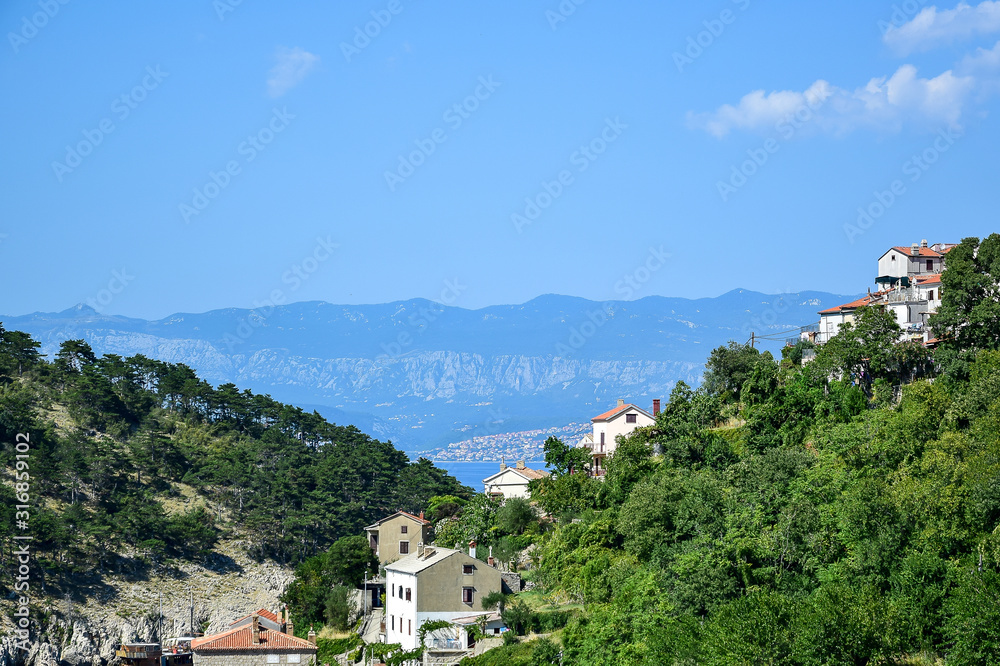 Vrbnik / Krk, Croatia - Panorama of the city, white and beige houses with orange roofs, green trees, blue sky with clouds, mountains and the sea in the background.