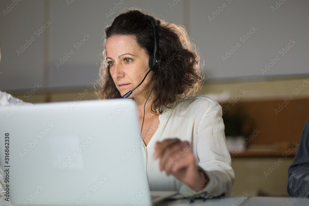 Focused mature call center operator looking at laptop. Serious curly woman working in office. Call center concept Focused mature call center operator looking at laptop. Serious curly woman working in office. Call center concept