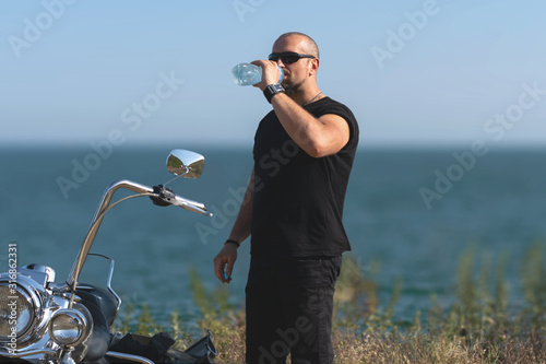 hot biker is drinking water on the motorcycle. Brutal man in glasses against the bike