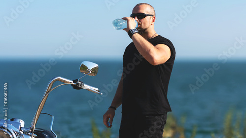 hot biker is drinking water on the motorcycle. Brutal man in glasses against the bike