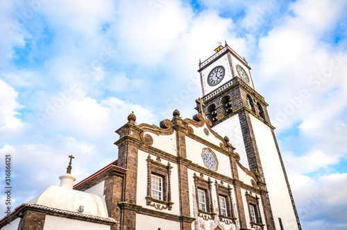 The outdoor facade of St. Sebastian Church, Igreja Matriz de Sao Sebastiao, in Ponta Delgada, Azores, Portugal. White clock tower from below with blue sky and clouds above. Sunset light, golden hour