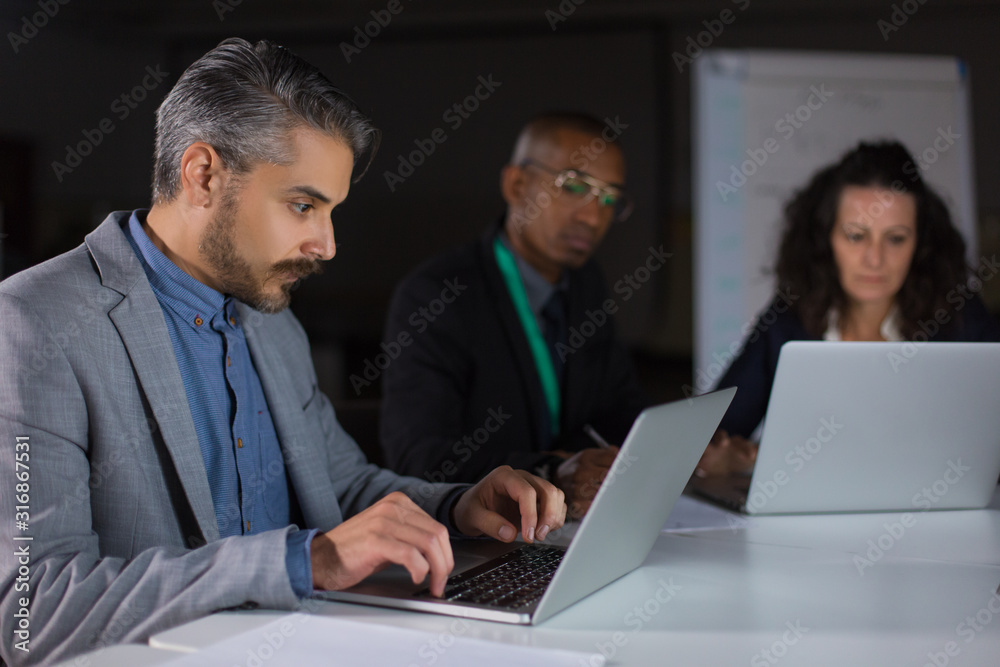 Focused man typing on laptop while working in office at night. Concentrated handsome manager sitting at table and looking at laptop. Business, working late concept Focused man typing on laptop while working in office at night. Concentrated handsome manager sitting at table and looking at laptop. Business, working late concept