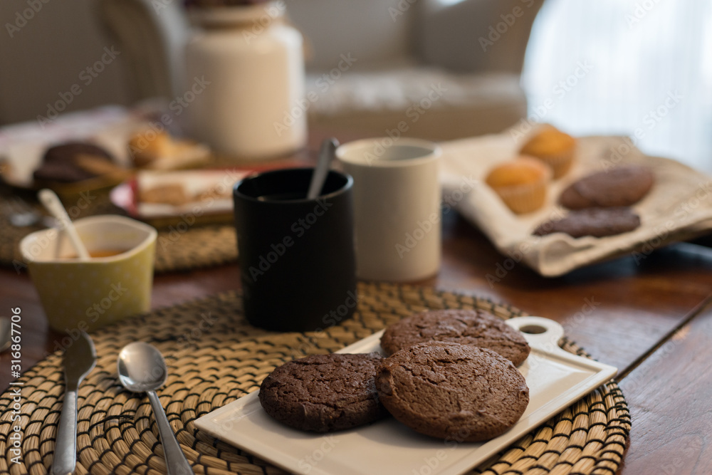 A rustic food set with homemade chocolate biscuits on a tray next to some mugs, cookies and muffins in a defocused background A rustic food set with homemade chocolate biscuits on a tray next to some mugs, cookies and muffins in a defocused background