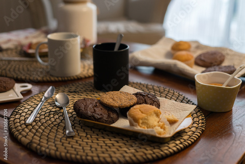 A rustic food set with a muffin in bits, homemade biscuits on a tray next to some mugs, cookies and sweets in a defocused background