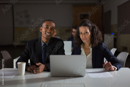 Smiling business partners looking at camera. Front view of cheerful employees sitting at table with laptop. Business, working late concept