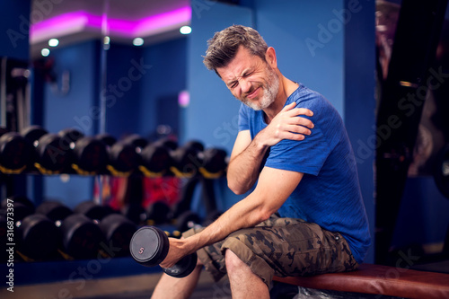Fototapeta Naklejka Na Ścianę i Meble -  Man feeling strong shoulder pain while training with dumbbells in the gym. People, fitness and healthcare concept