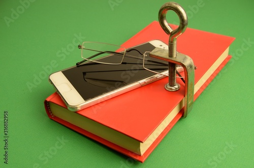 la lettura dei libri è stata sostituita dalla connessione dello smartphone ad Internet : libro chiuso da una morsa, uno smartphone  e un paio di occhiali photo