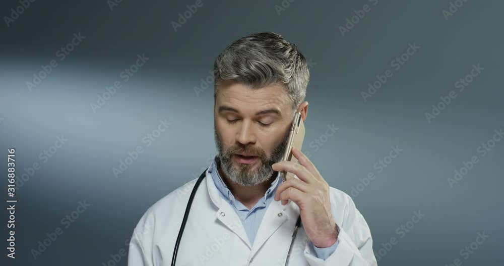 Caucasian good looking grey-haired male medic in white gown speaking on the mobile phone on the gray background. Close up. Caucasian good looking grey-haired male medic in white gown speaking on the mobile phone on the gray background. Close up.