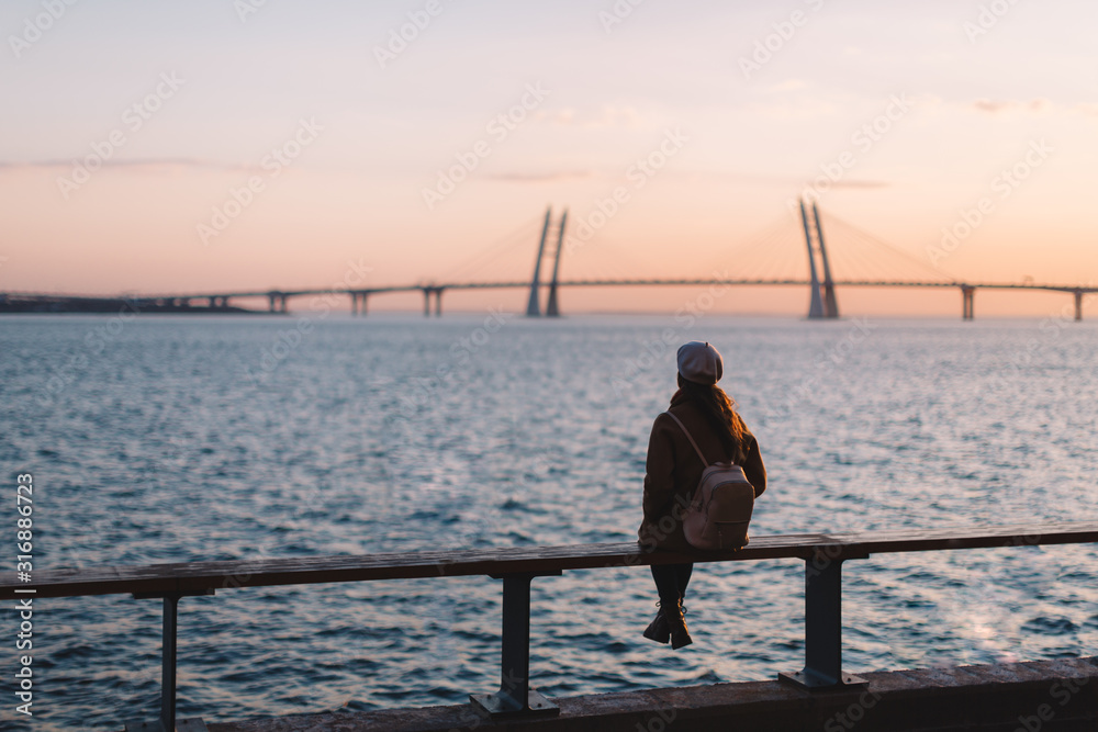 Alone dreamy unrecognizable girl in beige coat, white beret and with creamy backpack sitting on the edge of the sea. Female watching at colorful sunset. View from the back. Scenic portrait. Copy space Alone dreamy unrecognizable girl in beige coat, white beret and with creamy backpack sitting on the edge of the sea. Female watching at colorful sunset. View from the back. Scenic portrait. Copy space