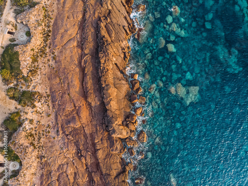 Above aerial view of rocky shore of Sardinia island in Mediterranean sea. Waves crash against rocks and reefs. Colorful blue color of sea water. Sunset time. Vacation and tourism concept.
