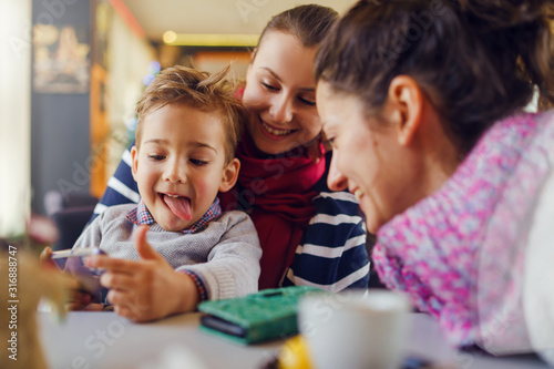 Small little boy caucasian child son sitting in the lap of his mother or aunt woman while female friend is looking to the smart mobile phone playing games or making a video call