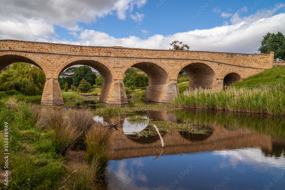 Richmond, Tasmania, Australia - December 13, 2009: Aong water line closeup shot of brown stone historic bridge over coal river reflected in water with reed and green lawn on side. Blue cloudscape.