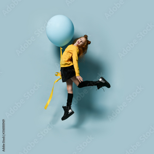 Teenager in yellow sweatshirt, black skirt, knee-highs, boots. She smiling, holding balloon, jumping up against blue background.