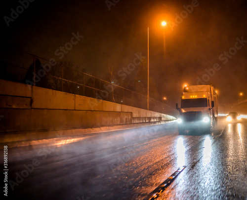 Big rig professional semi truck with semi trailer running on the wet rainy road at night time with turned on headlights