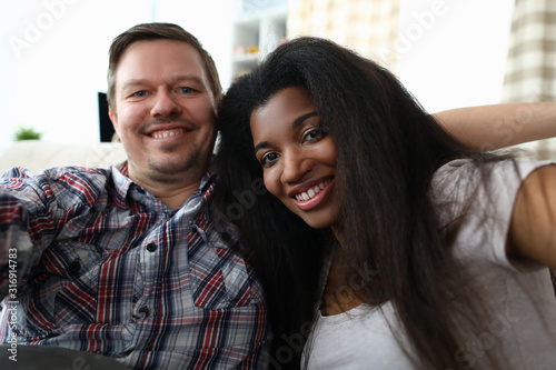 Portrait of cheerful mixed race couple taking happy selfie on sofa indoors. Man and latino woman widely smiling on camera. Multinational and biracial family concept