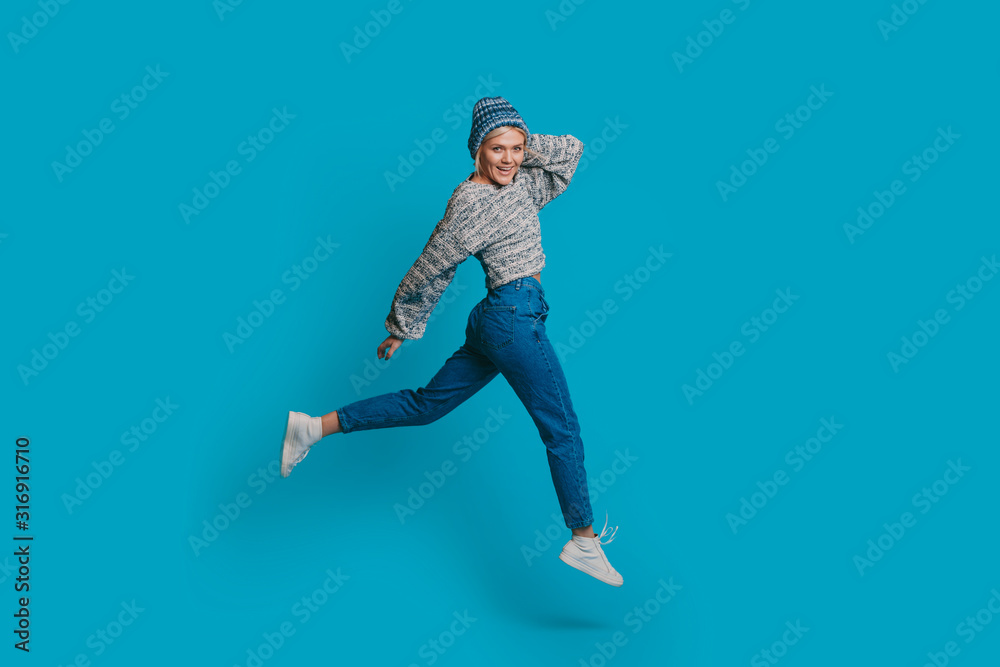 Full length portrait of a beautiful young blonde woman jumping while looking at camera smiling touching her head with a hand dressed in blue against a blue wall