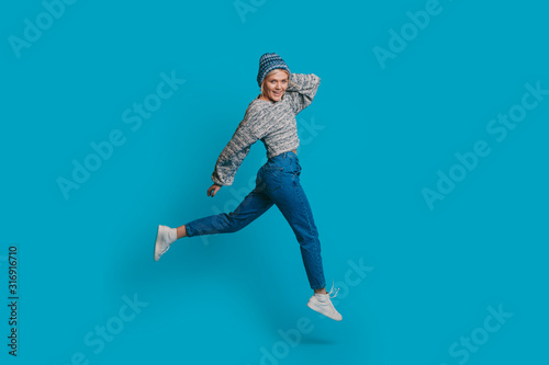 Full length portrait of a beautiful young blonde woman jumping while looking at camera smiling touching her head with a hand dressed in blue against a blue wall © Strelciuc
