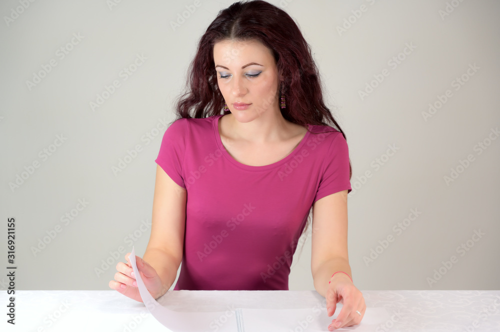 The model smiles and makes a financial report. Portrait of a pretty slender brunette woman in a pink dress and with brown hair sitting at a table on a white background in the studio. The model smiles and makes a financial report. Portrait of a pretty slender brunette woman in a pink dress and with brown hair sitting at a table on a white background in the studio.