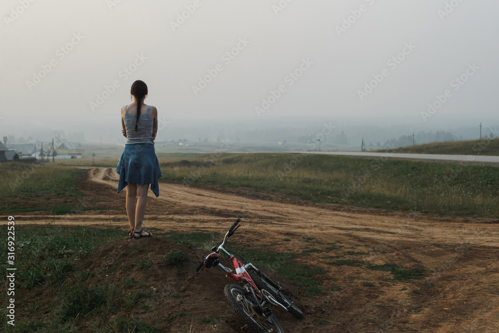 white caucasian young woman standing in village hill road with bike, looking at open space, view from back in full body size, horizontal lifestyles stock photo image with copy space for text