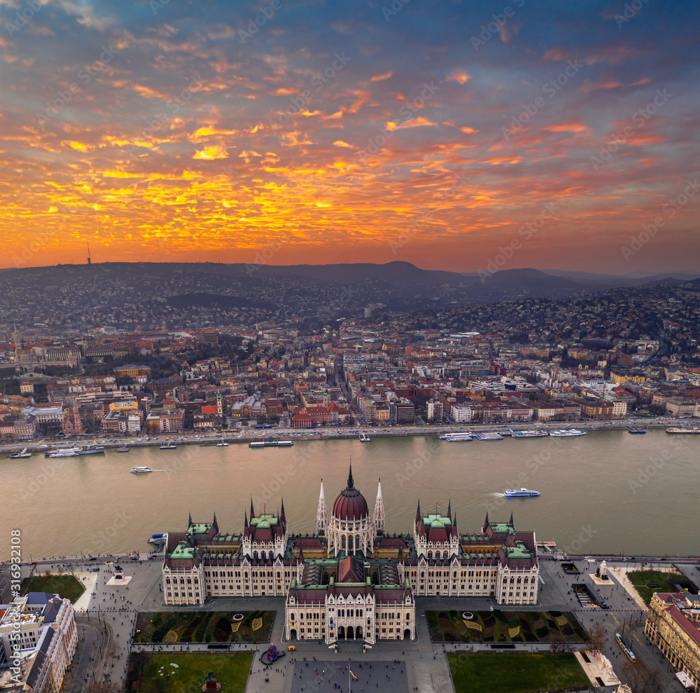Budapest, Hungary - Aerial panoramic drone view of the Parliament of Hungary at sunset with beautiful dramatic golden clouds, Christmas tree and sightseeing boats on River Danube
