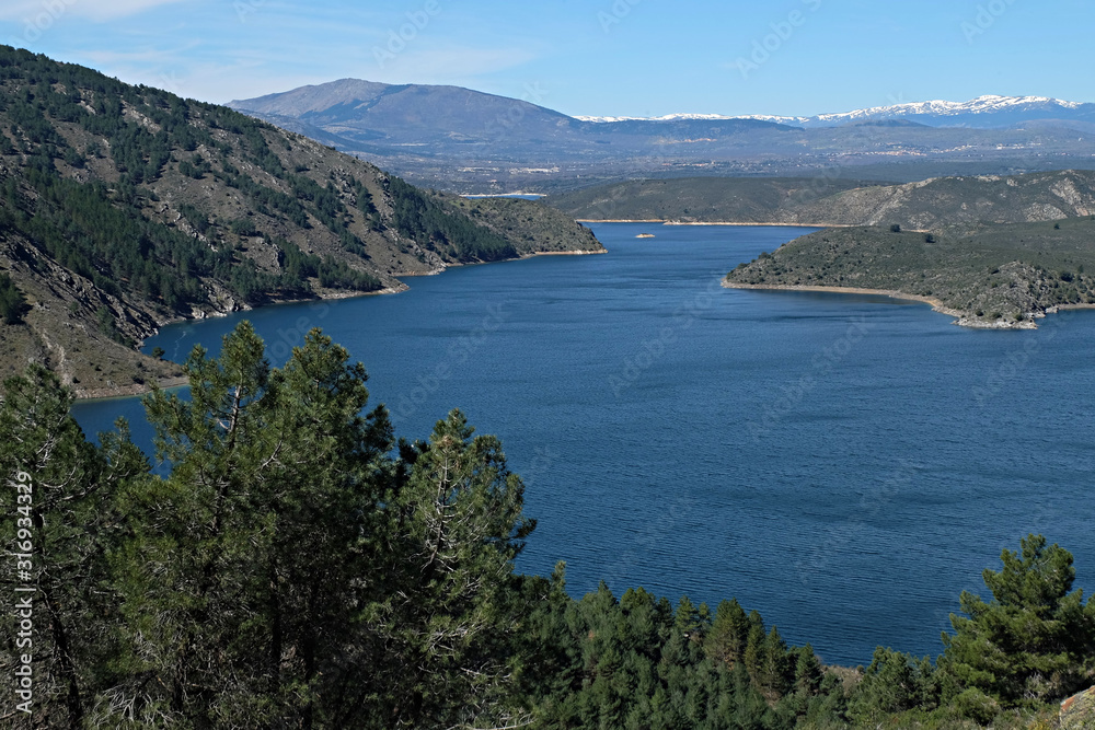 Natural landscape with reservoir and trees in the foreground, mountains in the background and intense blue sky, El Atazar reservoir, Madrid, Spain