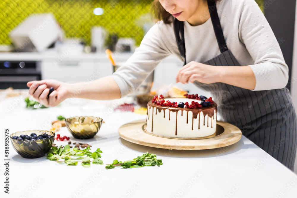 Confectioner decorates with berries a biscuit cake with white cream and chocolate. Cake stands on a wooden stand on a white table. The concept of homemade pastry, cooking cakes. Confectioner decorates with berries a biscuit cake with white cream and chocolate. Cake stands on a wooden stand on a white table. The concept of homemade pastry, cooking cakes.