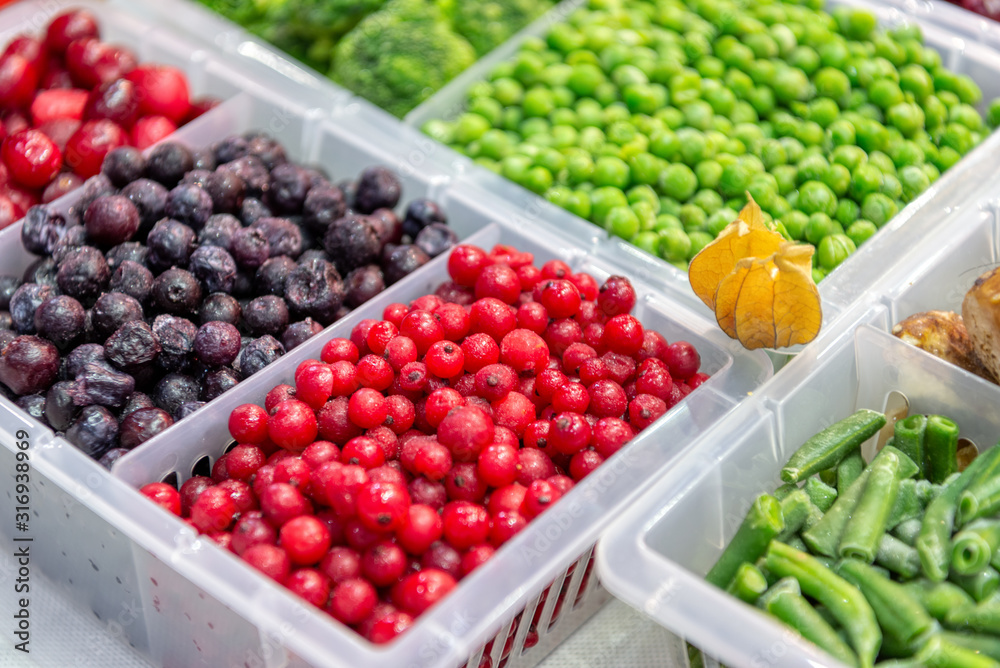 Frozen fruits and vegetables. Products are poured into plastic boxes. In the center of the frame is red and black currants. Freezer shop window.