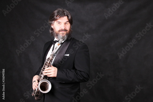 Fotografia A male artist musician in a classic black suit, tailcoat, statuesque in a bow tie with a beard plays music on a gold saxophone