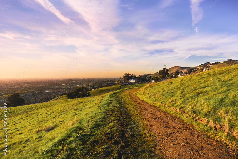 Obraz premium Sunset view of hiking trail on the verdant hills of East San Francisco Bay Area; the City of Hayward visible in the background; California