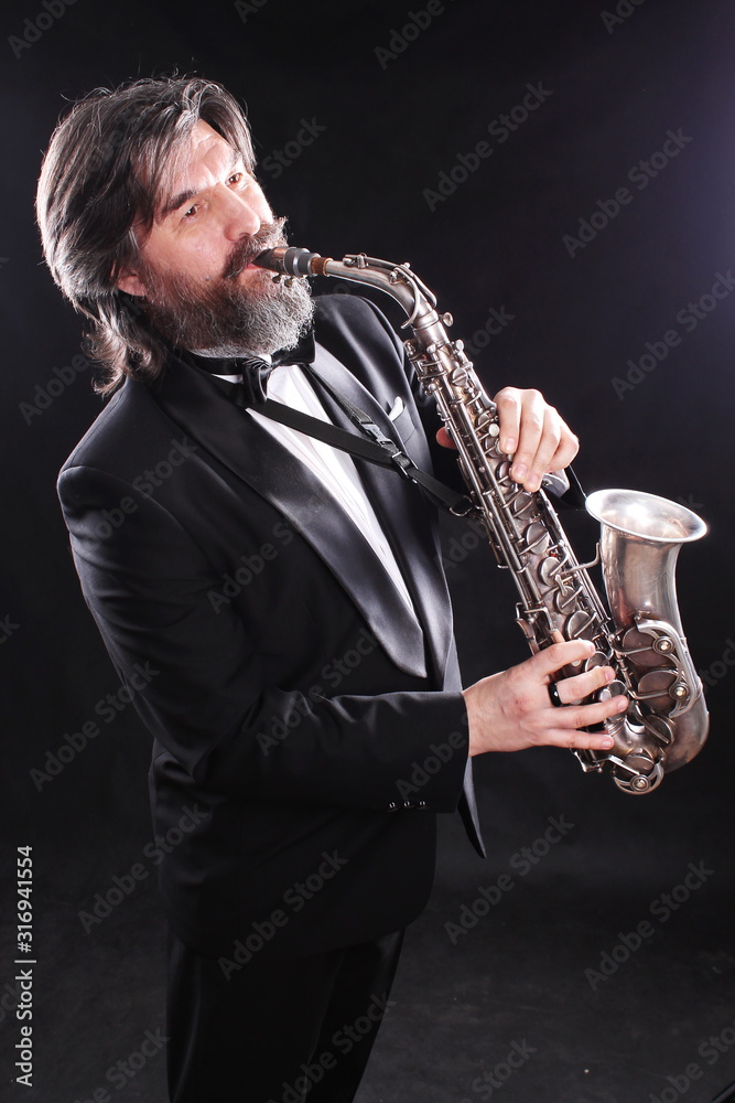 Naklejka premium A male artist musician in a classic black suit, tailcoat, statuesque in a bow tie with a beard plays music on a gold saxophone.Playing an instrument against a white light source.black background