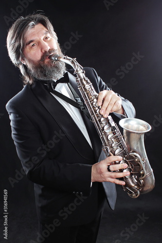 Fototapeta A male artist musician in a classic black suit, tailcoat, statuesque in a bow tie with a beard plays music on a gold saxophone