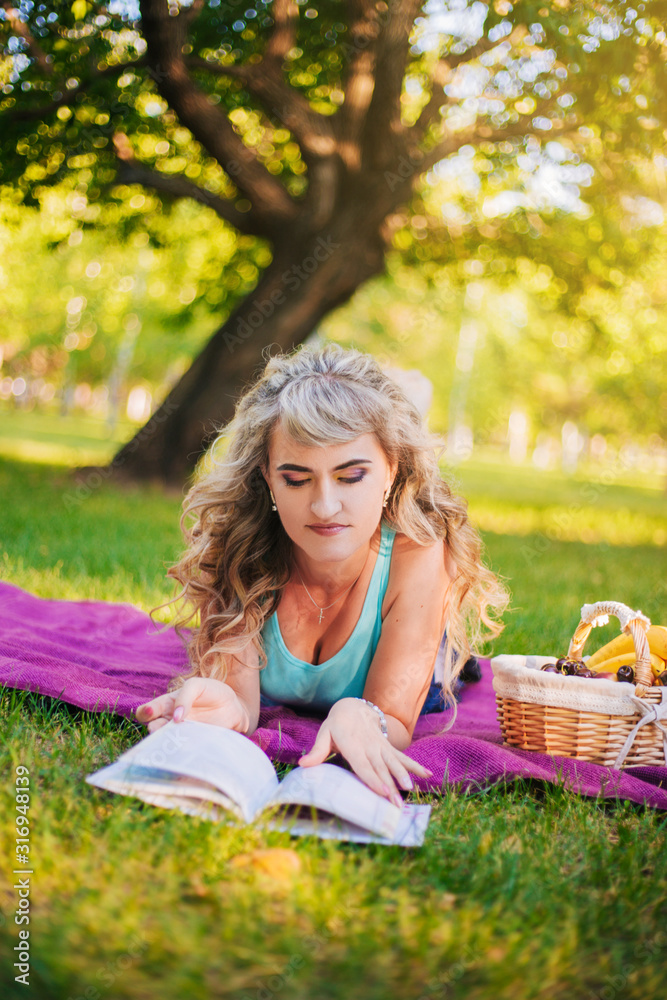 Beautiful girl on a picnic in the park is reading a book. Blonde on a purple bedspread resting. Fruit basket at a picnic Beautiful girl on a picnic in the park is reading a book. Blonde on a purple bedspread resting. Fruit basket at a picnic