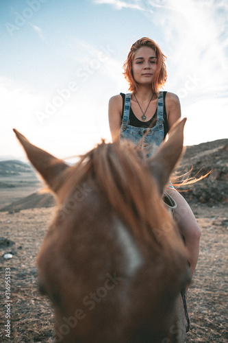 a young girl with red hair sees riding a red horse at sunset along a hill scorched by the sun against a background of high snow peaks