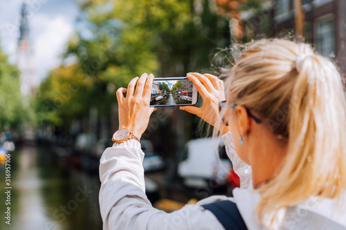 Blonde Woman tourist taking a photo of famous Soutern church (Zuiderkerk) and canal in Amsterdam on the mobile phone. Warm gold afternoon sunlight. Travel in Europe, city trip concept. photo