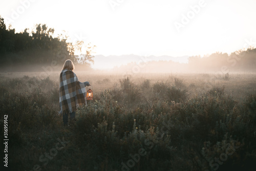 young girl with blond hair in a warm woolen sweater wrapped in a plaid, she is standing in a field with tall dry grass at dawn with a kerasin lamp in her hands