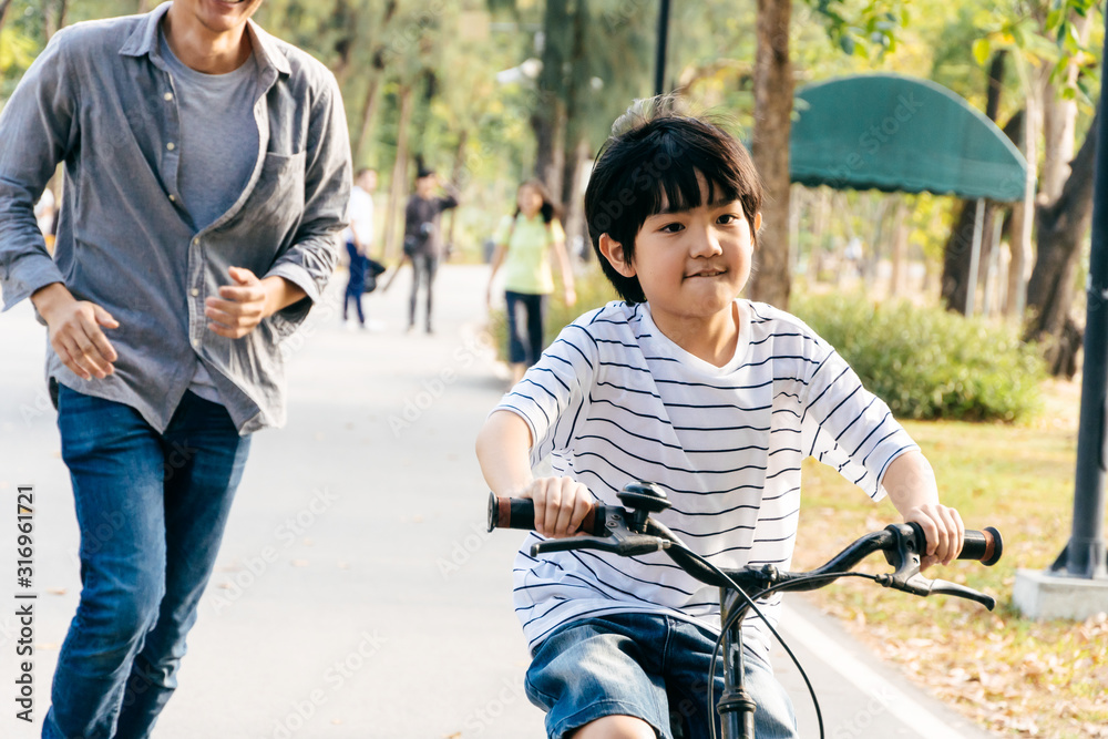 Happy Asian boy riding a bicycle with his father running behind to keep his kid safe and support at park, Dad teach son lesson and spend time together on weekend concept - obrazy, fototapety, plakaty 