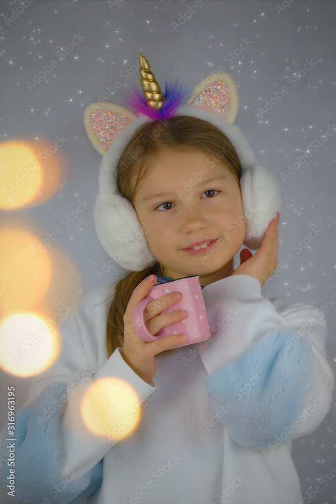Cute girl has warm plush ear muffs with horn unicorn. Holding pink mug with polka dots.