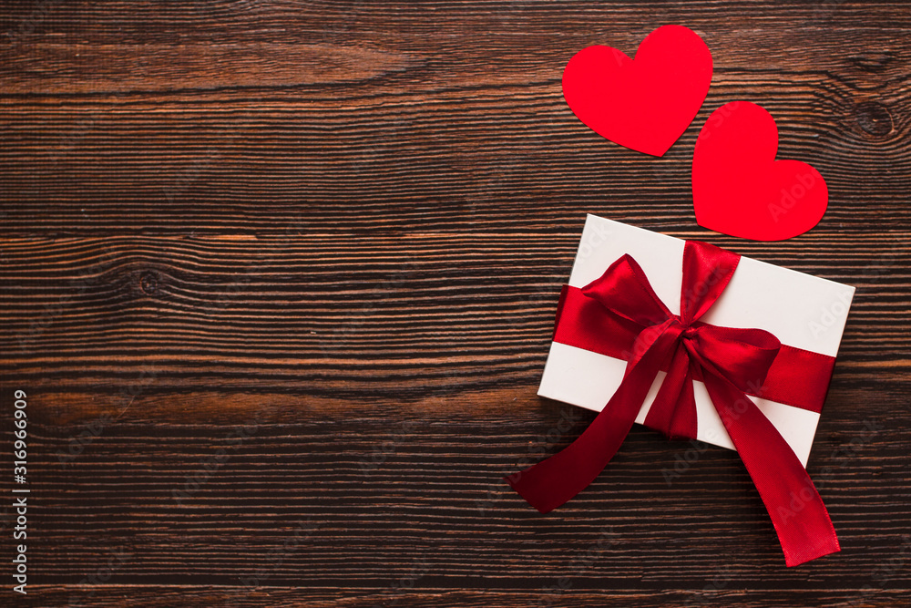 White present with a red ribbon and paper hearts isolated on a dark wooden background. Top side view of a celebrating warm flatlay. Valentines day and Christmas concept. Copyspace. White present with a red ribbon and paper hearts isolated on a dark wooden background. Top side view of a celebrating warm flatlay. Valentines day and Christmas concept. Copyspace.