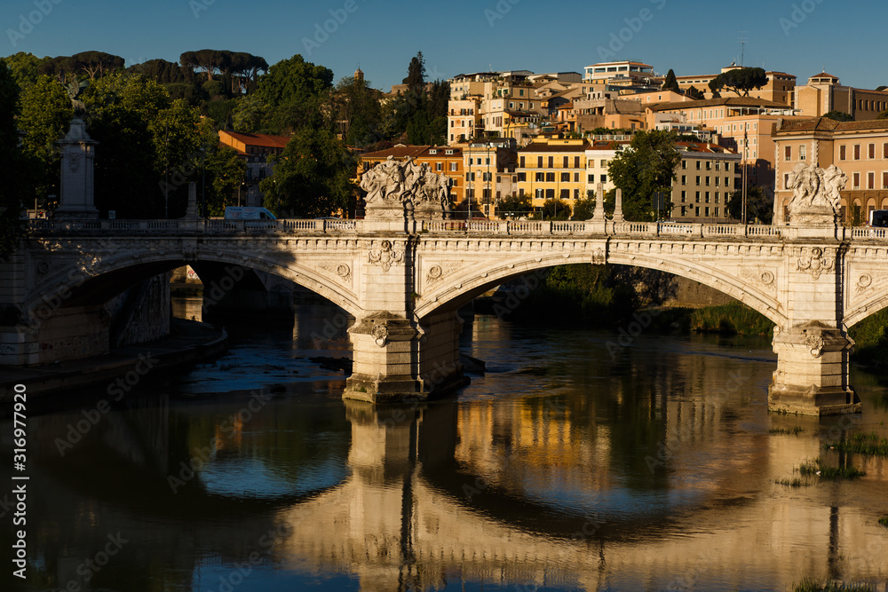 The beautiful and majestic eternal city of Rome, the capital of Italy, in the early morning at sunrise from the bridge of San Angelo, the view of the bridge and the Tiber river and the dawn reflection