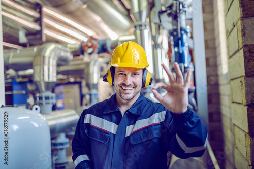 Fotografering Smiling satisfied blue collar worker in working clothes, with helmet and hardhat standing in factory and showing okay sign