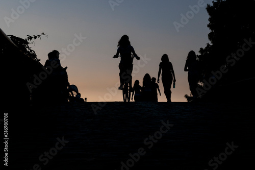 People silhouettes seen in a park on top of an arched footbridge in various poses.